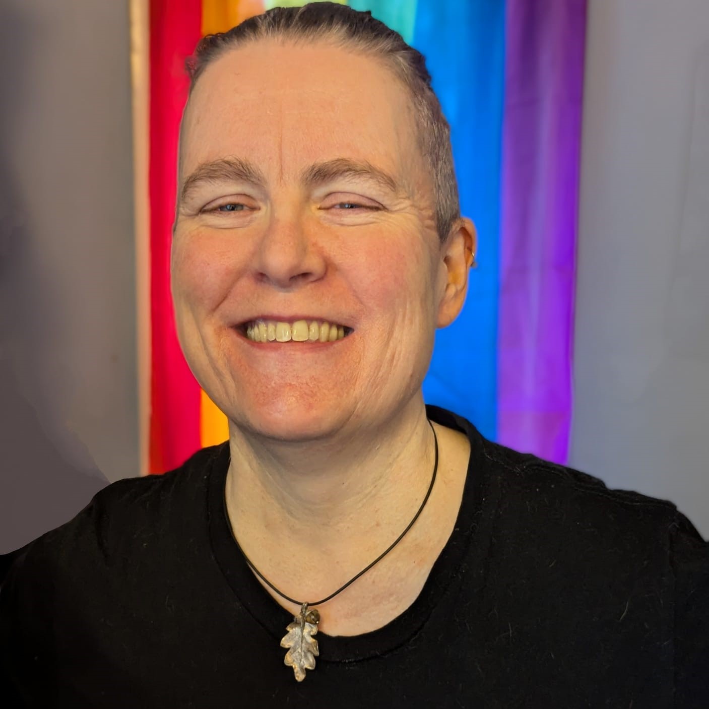Photo of Lucy, SShort hair, black shirt, big smile and a necklace. LGBTQ flag in backdrop